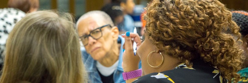 Photo of an African American woman speaking as another African American women listens intently